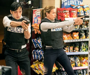 Police officers with guns drawn in a convenience store aisle, wearing tactical vests labeled "POLICE".