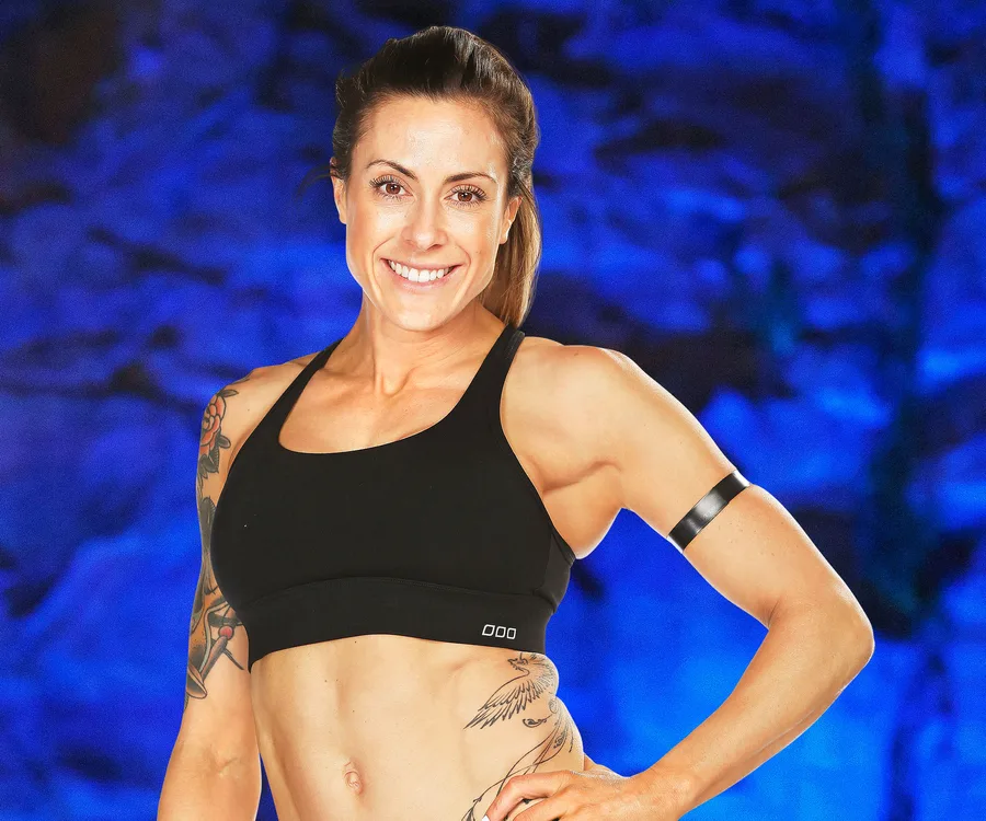 A fit woman with tattoos wearing a black sports bra, smiling confidently against a blue abstract background.