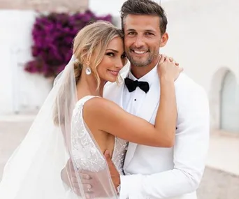 A couple in wedding attire smiling, the bride wearing a veil and the groom in a tuxedo, embracing outdoors.