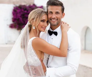 A couple in wedding attire smiling, the bride wearing a veil and the groom in a tuxedo, embracing outdoors.