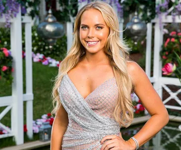 Blonde woman in sparkly silver gown smiling outdoors with flowers and lights in background.
