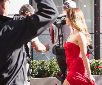A woman in a red dress walks on a set with crew members and petals in the air.