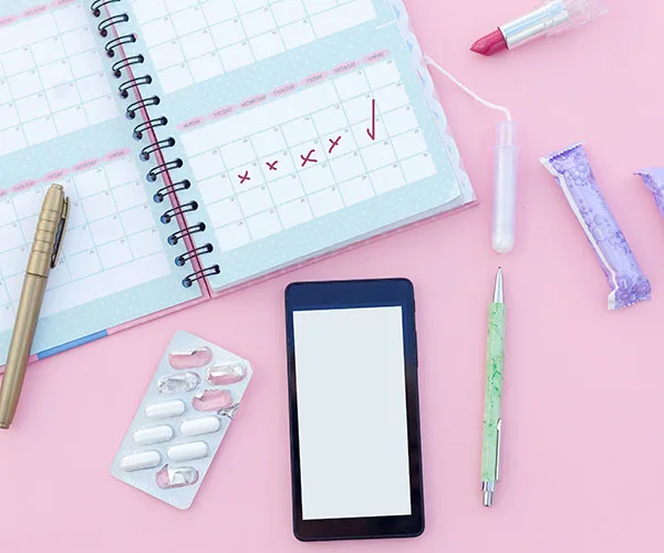 Calendar with marked dates, blister pack of pills, smartphone, pens, tampons, and lipstick on a pink background.
