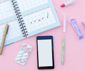 Calendar with marked dates, blister pack of pills, smartphone, pens, tampons, and lipstick on a pink background.