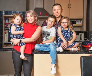 A family of five in a kitchen, parents with three smiling children, two girls in matching dresses and a boy in jeans.