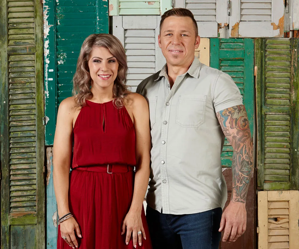Two people standing in front of rustic shutters, with one wearing a red dress and the other in a light shirt showing tattoos.