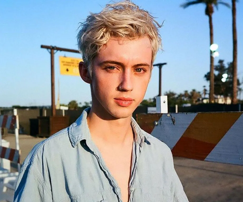 Young person with short blonde hair in a light blue shirt, standing outdoors beside a road barrier, under a clear sky.