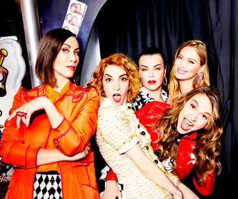 Five women pose in vibrant outfits in a photo booth setting with a playful and energetic vibe.