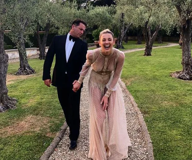 A couple dressed formally, laughing and holding hands, walk through a garden path lined with trees.