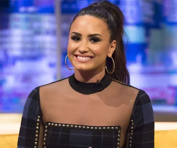 Demi Lovato smiling during a TV interview, wearing a black and sheer top with hoop earrings.