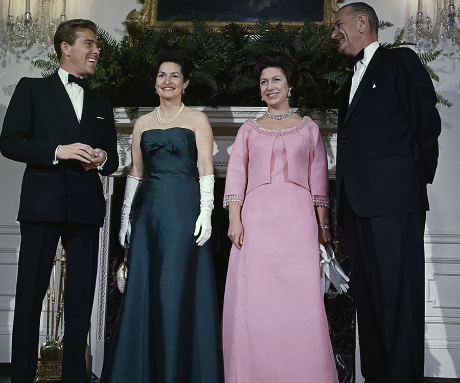 A formal event with two women in elegant gowns and two men in tuxedos, standing in front of a fireplace with greenery.
