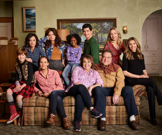 A group photo of "The Conners" cast, sitting and standing around a couch in a living room setting.