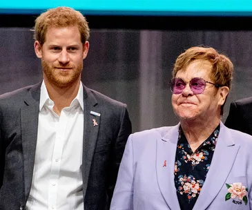 Prince Harry and Elton John together at an AIDS awareness event, both wearing suit jackets with red ribbon pins.