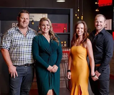 Group of four people posing indoors, two men and two women, dressed in semi-formal attire, standing in a well-lit room.