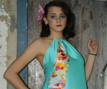 A person in a sleeveless turquoise dress with floral accents, standing in front of a rustic wooden door.