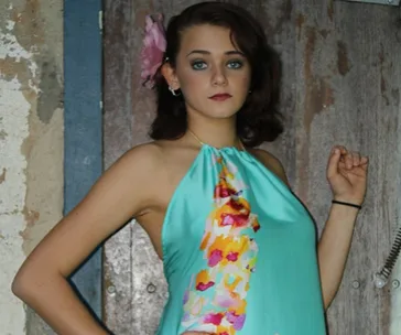 A person in a sleeveless turquoise dress with floral accents, standing in front of a rustic wooden door.