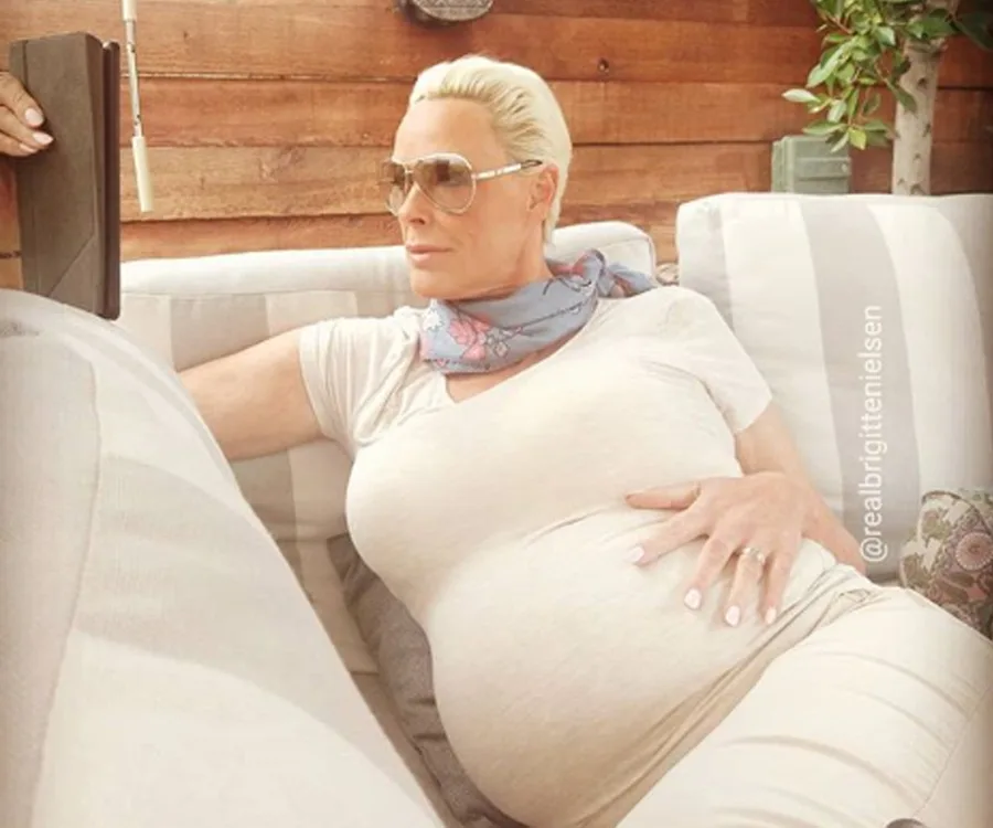 Pregnant woman relaxing on a couch, wearing sunglasses and scarf, hand on belly. Sunlit outdoor setting.