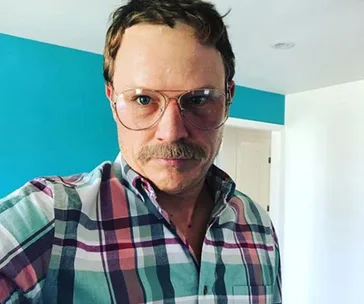 Man with a mustache, wearing glasses and a plaid shirt, stands indoors against a blue wall.
