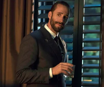 A man in a suit holding a glass, standing by window shutters, looking contemplative.