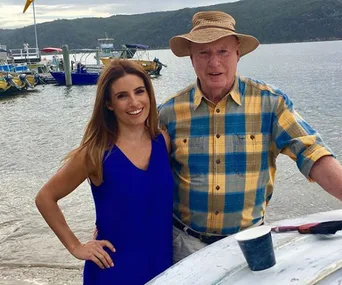 Man in flannel shirt and hat stands with woman in blue dress by a boat at a lakeside with hills in the background.