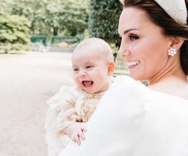A woman smiling at a baby in her arms, outdoors with trees in the background.
