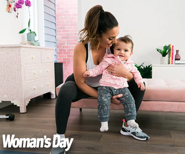 Woman workouts with baby in living room, smiling, wearing gym clothes, baby in pink jacket. "Woman's Day" logo in corner.