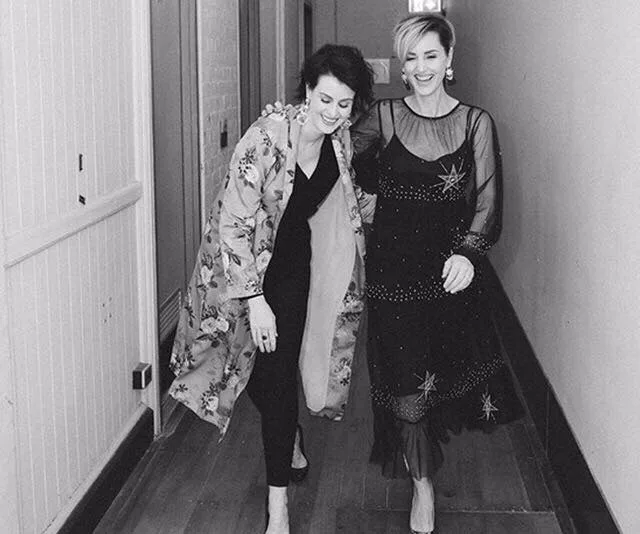 Two women laughing in a hallway, one wearing a floral coat, the other in a sheer star-patterned dress.