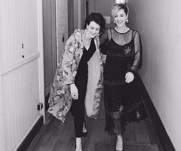 Two women laughing in a hallway, one wearing a floral coat, the other in a sheer star-patterned dress.