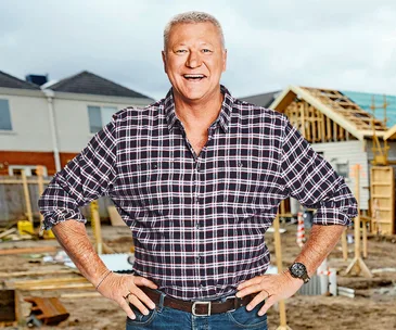 Man in plaid shirt stands smiling at a construction site with partially built houses in the background.