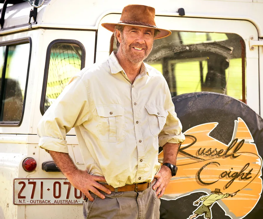 Man in khaki attire and hat stands smiling beside an off-road vehicle with 'Russell Coight' logo in the background.