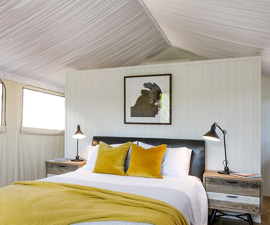 Glamping tent interior with a cozy bed, yellow pillows, and artwork of a black cockatoo on the wall.