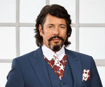 Laurence Llewelyn-Bowen in a blue suit with a patterned tie and pocket square, standing in front of a window.
