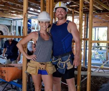 A smiling couple in construction gear stands at a building site.