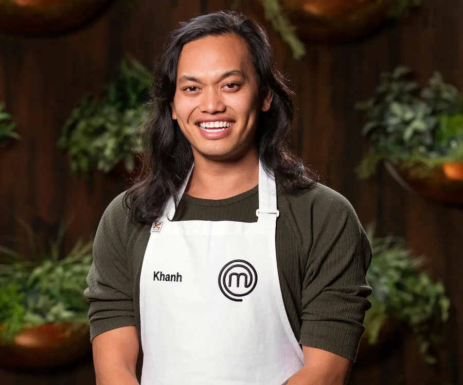 Khanh Ong smiling in a MasterChef apron, standing in front of a dark background with greenery.