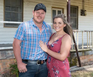 Couple smiling in front of a rustic house; man in a plaid shirt and cap, woman in a floral dress.