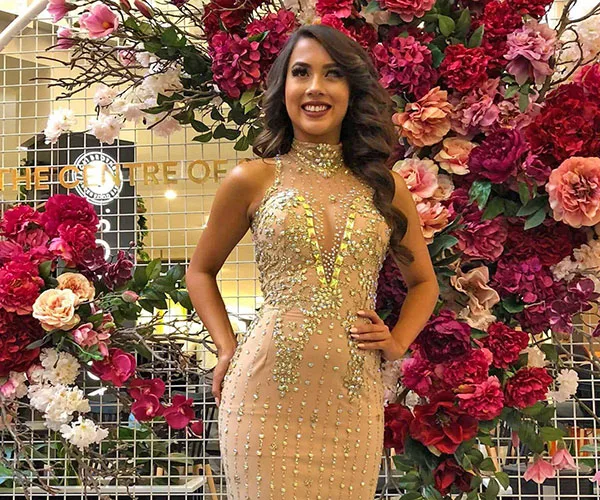 Woman in a glittering gown stands confidently surrounded by vibrant floral backdrop.