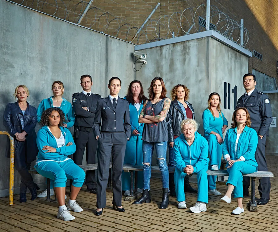 Cast of "Wentworth" in prison yard, characters in uniforms and casual wear, standing and seated with barbed wire background.