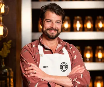 Ben, a MasterChef Australia 2018 runner-up, smiling in a patterned shirt and apron, standing in a warmly lit room.