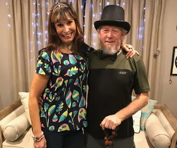 A woman in a colorful shirt and tiara hugs an older man in a top hat, standing in a cozy room with soft lighting.