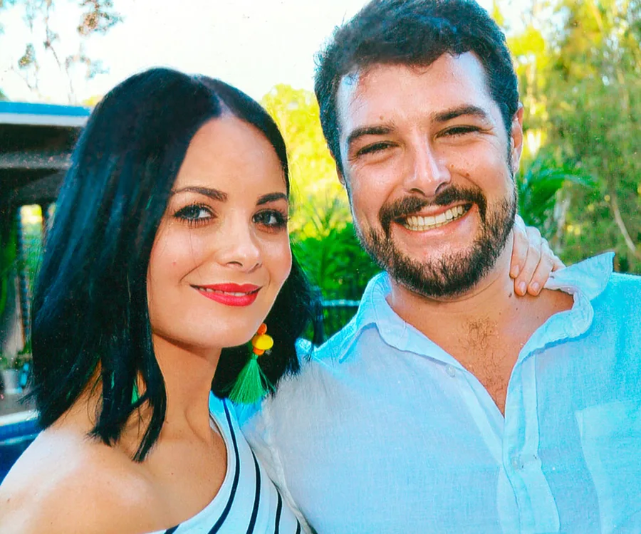 A smiling couple posing outdoors with trees in the background, the woman has black hair and the man a beard.