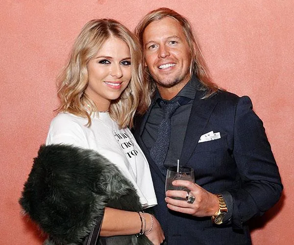 A couple, smiling, in formal attire against an orange background at an event.
