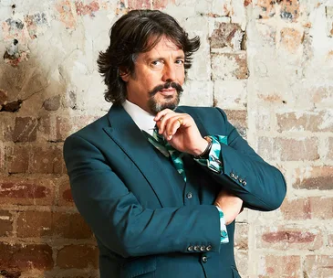 A man in a teal suit stands against a brick wall, looking thoughtful and confident.