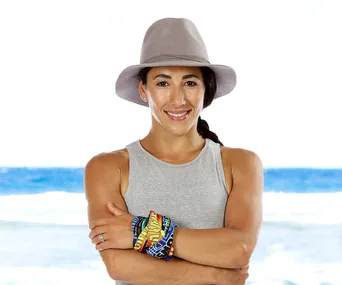 Woman in a gray tank top and hat, smiling on a beach, with colorful Survivor buffs on arms.