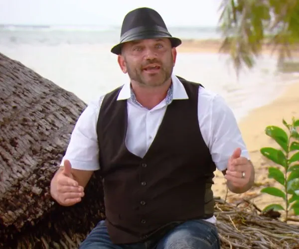 Man in fedora and vest sitting on beach, gesturing with hands, with ocean and tree in background.