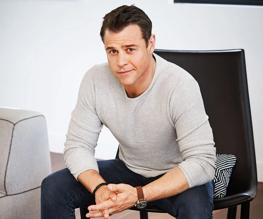 A person in a light sweater and jeans sits on a chair, hands clasped, looking at the camera.