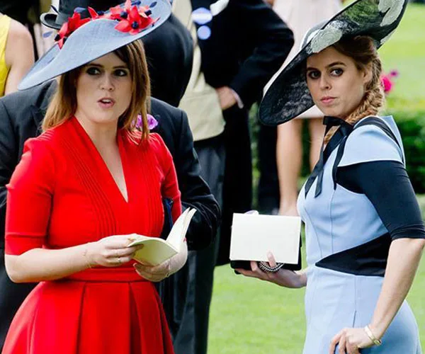 Two women in formal dresses and hats at an outdoor event, holding papers.