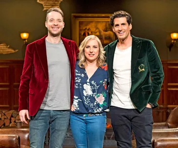 Three people standing together, two wearing colored jackets, in a warmly lit room with a painting in the background.