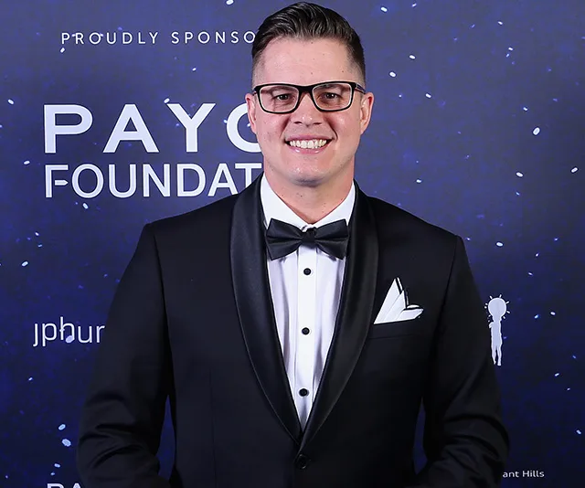 Man in tuxedo and glasses smiling at a formal event with a navy blue backdrop featuring text and logos.