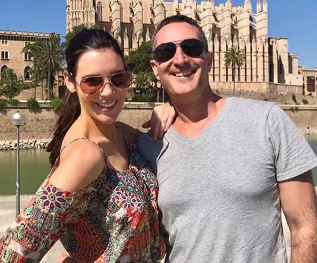 Two people smiling in front of a historic building, both wearing sunglasses, with water and palm trees in the background.