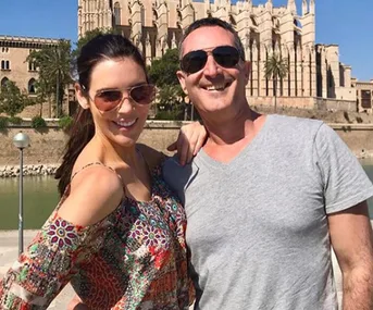 Two people smiling in front of a historic building, both wearing sunglasses, with water and palm trees in the background.